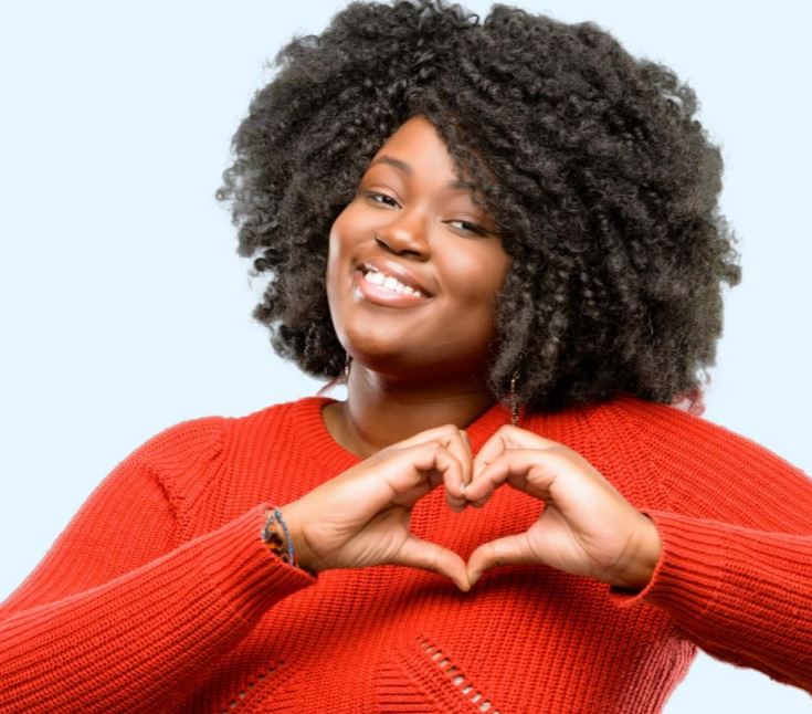 A Black woman wearing red is smiling and making the heart sign with her hands.
